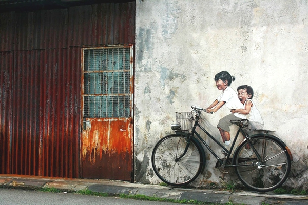 Observamos una vieja puerta metalica oxidada y al lado una pared de cemento donde hay una vieja bicicleta y pintaron sobre la pared unos niños que simulan estar montados sobre ella