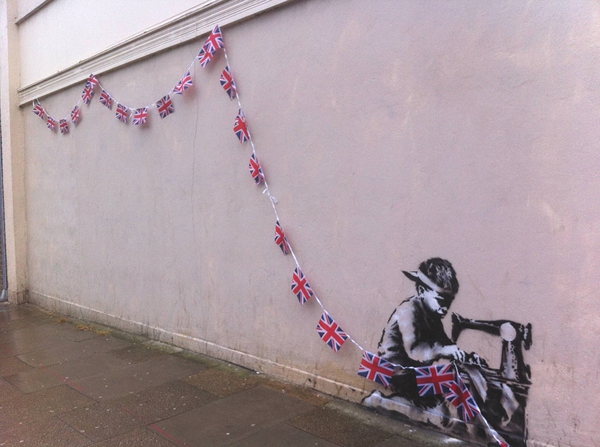 Vemos una pared de cemento donde se hallan unas banderas de Londres y al final de la pared un chico pintado con una maquina de coser que hace las banderas             