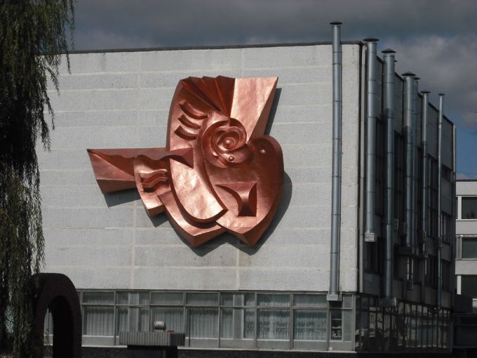 Vemos una edificación donde hay una escultura pegada a la pared una paloma en su pico tiene un átomo