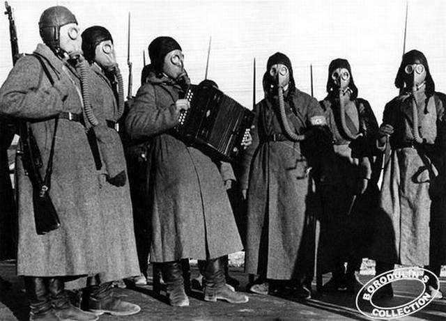 Vemos a seis personas usando mascaras anti-gases antiguas y abrigos  todos armados uno solo toca un instrumento musical 