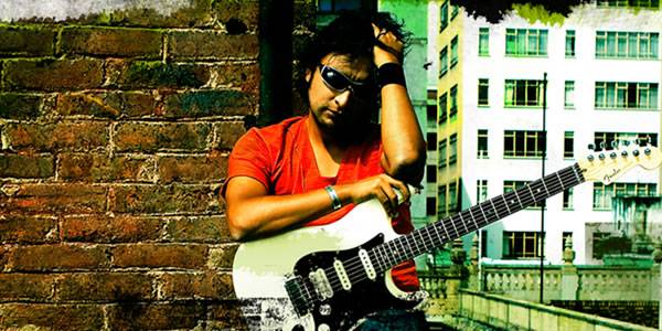 Un músico con su guitarra y gafas oscuras que toca su cabeza recostado en un muro de ladrillo