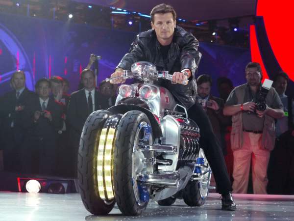 Vemos la moto con un conductor y sus luces prendidas personas tomandole fotos