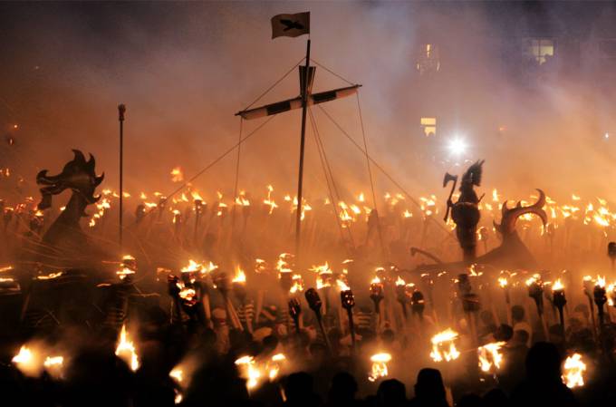 Podemos ver un gran barco vikingo y muchas antorchas en medio de la noche mientras que miles de personas con antorchas caminan a su alredor