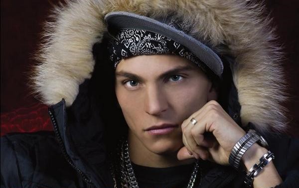 Un músico con una gorra y encima una capucha con piel y en sus brazos lleva anillo pulseras  y una cadena