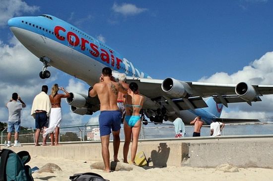Vemos un avion en colores blanco con azul que pasa sobre la playa casi rosando las cabezas de los turistas