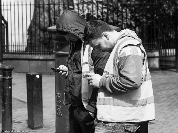 Vemos como dos hombres con chaquetas con capucha observan sus celulares en una calle