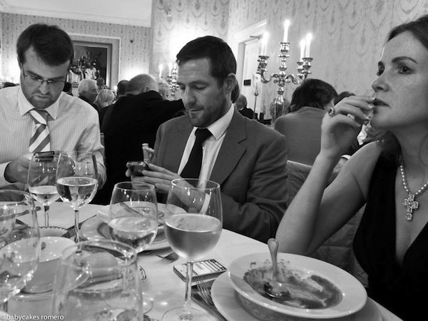 Tenemos en un elegante salón a varias personas disfrutando de una gran cena mientras todas observan sus celulares