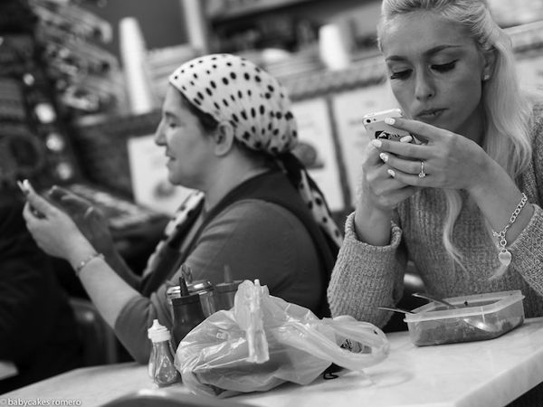 Vemos como dos mujeres comen  y las dos ven sus celulares una muy elegante la otra mas sencilla y con pañuelo atado a su cabeza