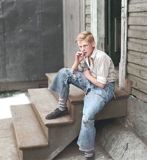 Joven rubio fumando con pantalón roto sentado en una escala de una casa misera