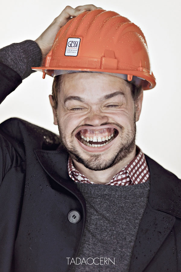 Un hombre con un  casco de protección sobre su cabeza lo sostiene con su mano sus ojos cerrados y su boca abierta y sus labios hacia arriba  muestran dientes y encias 