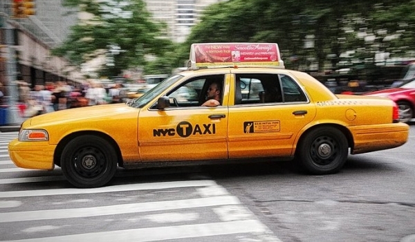 Tenemos un taxi amarillo que  lleva en su parte alta una publicidad de algo pasa por  un sitio con zona verde y edificios