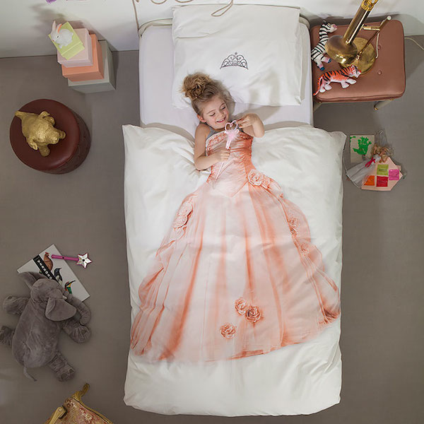 Un cama donde hay un edredon blanco que trae dibujado un hermoso traje largo para niña y donde la niña se cubre con el y parece que ella tuviera puesto el vestido