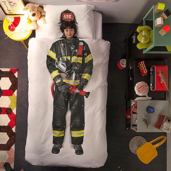 Vemos un edredón  que tiene pintado un traje de bombero y dentro un niño que parece lo llevase puesto  al lado de la cama hay una mesa donde se observa obetos propios del mundo bomberil