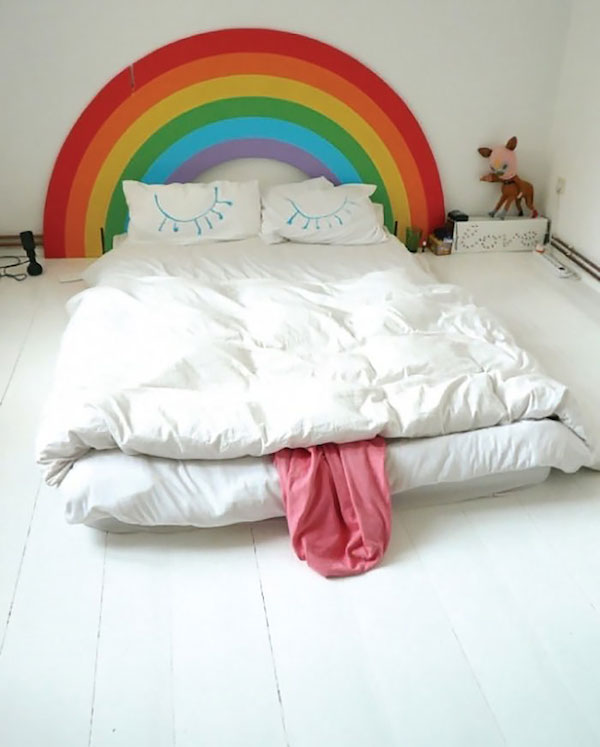 Vemos una cama grande donde el testero de la cama muestra un arco iris en colores fuertes y luego las dos almohadas muestran las pestañas  de un muñeco  