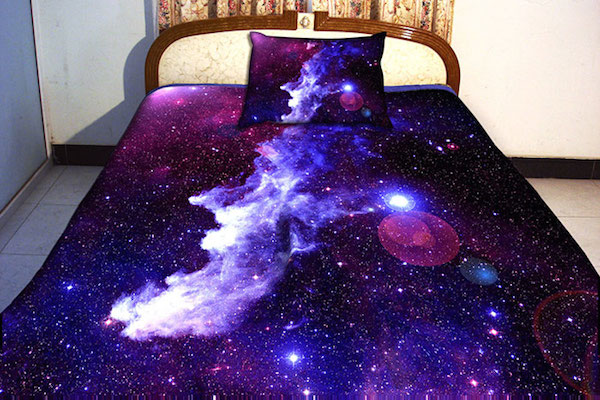 Vemos una cama con  un tendido que representa el universo plagado de estrellas y galaxias en colores morados y lilas y brillantes estrellas 